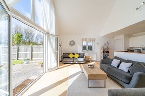 A spacious living area with a large sliding glass door, a grey sofa, a wooden coffee table, and a kitchen area in the background.