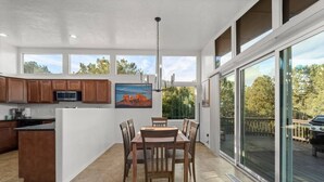 A modern kitchen with wooden cabinets, a dining area with a table and chairs, and large windows offering views of trees and a landscape painting. - Uptown Home w/ Amazing Views and Hot Tub (Sedona)