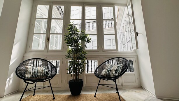 A room with two black wicker chairs, a potted plant, and a large window with white shutters. - Apto Plus 1 Dorm. Int. 1ª Planta (Cádiz)