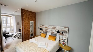 A modern bedroom with a wooden headboard, a bed with white linens and yellow pillows, a bedside table with a lamp, and a view of a balcony with a chair. - Apto Plus 1 Dorm. Int. 1ª Planta (Cádiz)