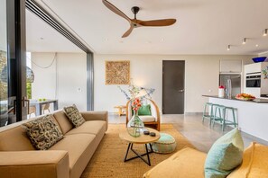 Una sala moderna con un sofá, una mesa de centro y una silla. Se observa un área de cocina con un refrigerador y una mesa de comedor.