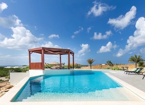 En swimmingpool med et træbeklædt pergola, liggestole og frit vue over havet.