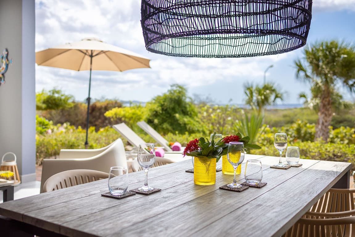 An outdoor dining area with a wooden table set for two, featuring yellow vases with flowers and glasses.