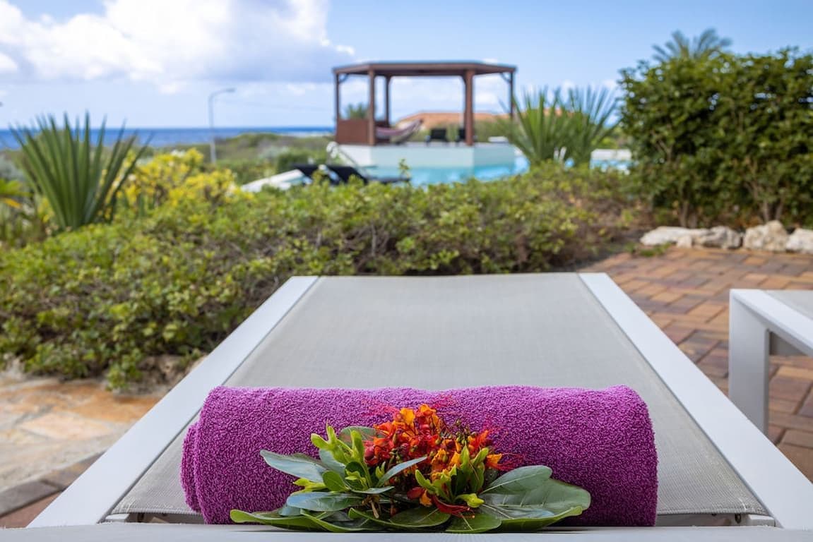 A lounge chair with a purple towel and a flower arrangement, overlooking a pool and a gazebo.