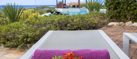 A lounge chair with a purple towel and a flower arrangement, overlooking a pool and a gazebo.
