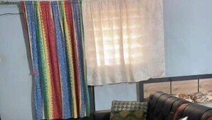 Un salón con un sofá negro y naranja, una manta a rayas colorida y una ventana con cortinas translúcidas.