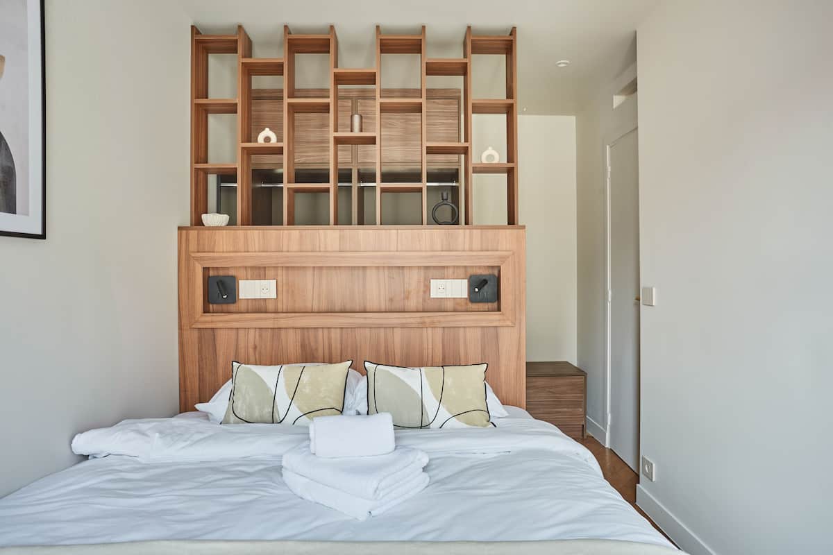 A neatly made bed with white linens and a folded towel, a wooden headboard with a built-in shelf, and a small bedside table with a lamp.