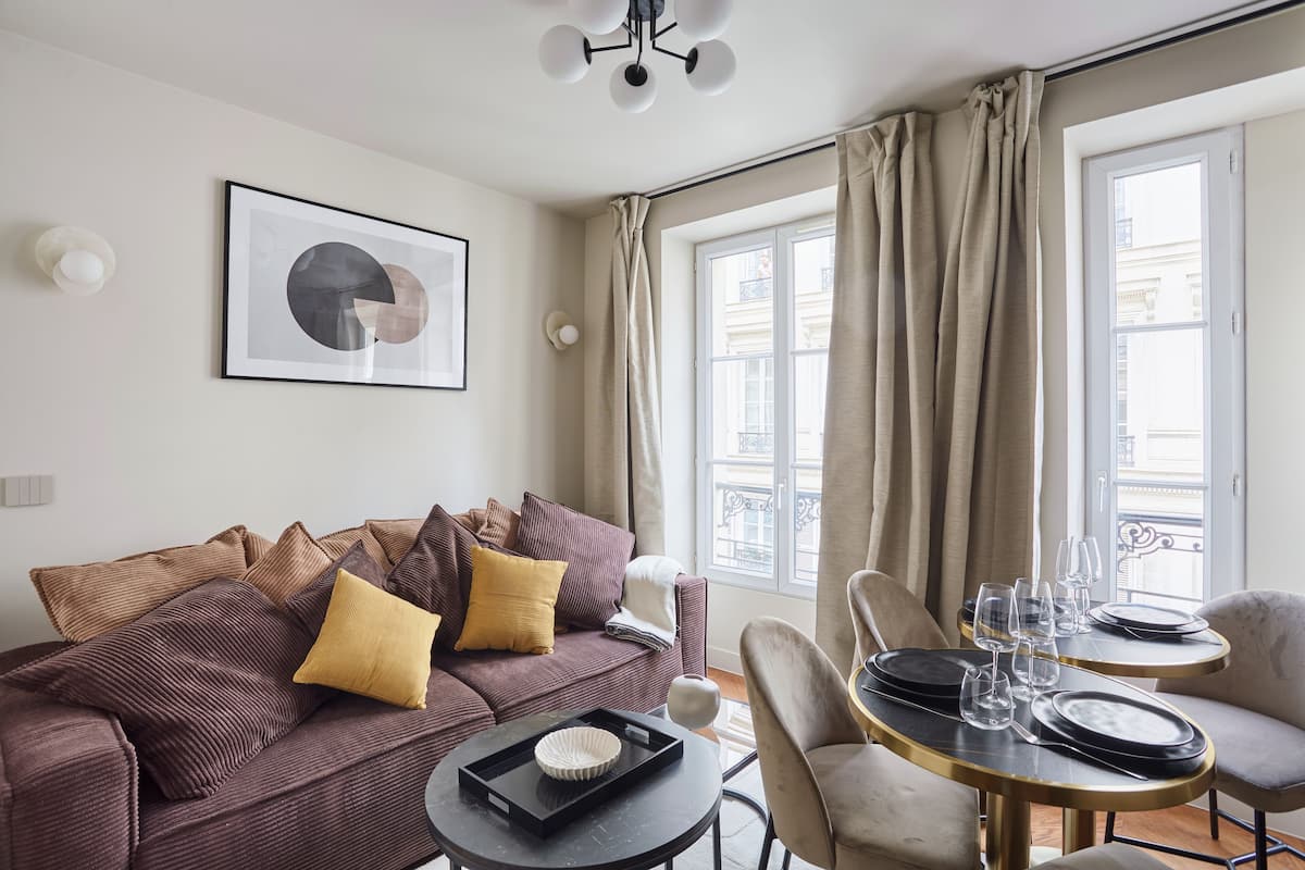 A modern living room with a sofa, a dining table set for two, and a large window with curtains.