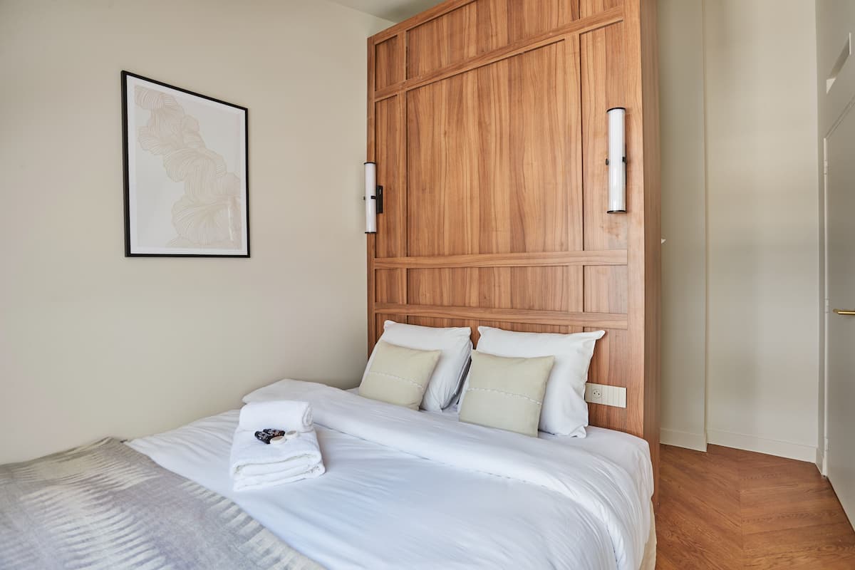 A neatly made bed with white linens, a wooden headboard, and a framed artwork on the wall.