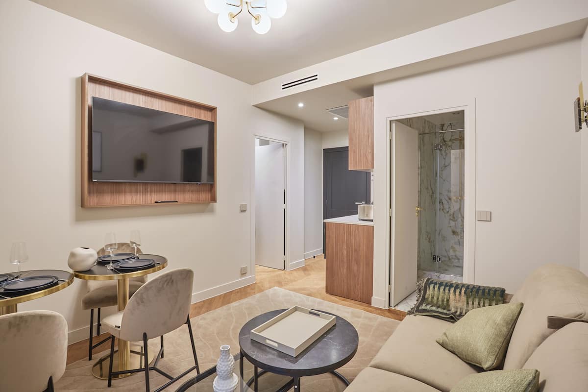 A modern living room with a flat-screen TV, a dining area with a round table and chairs, and a sofa with cushions.