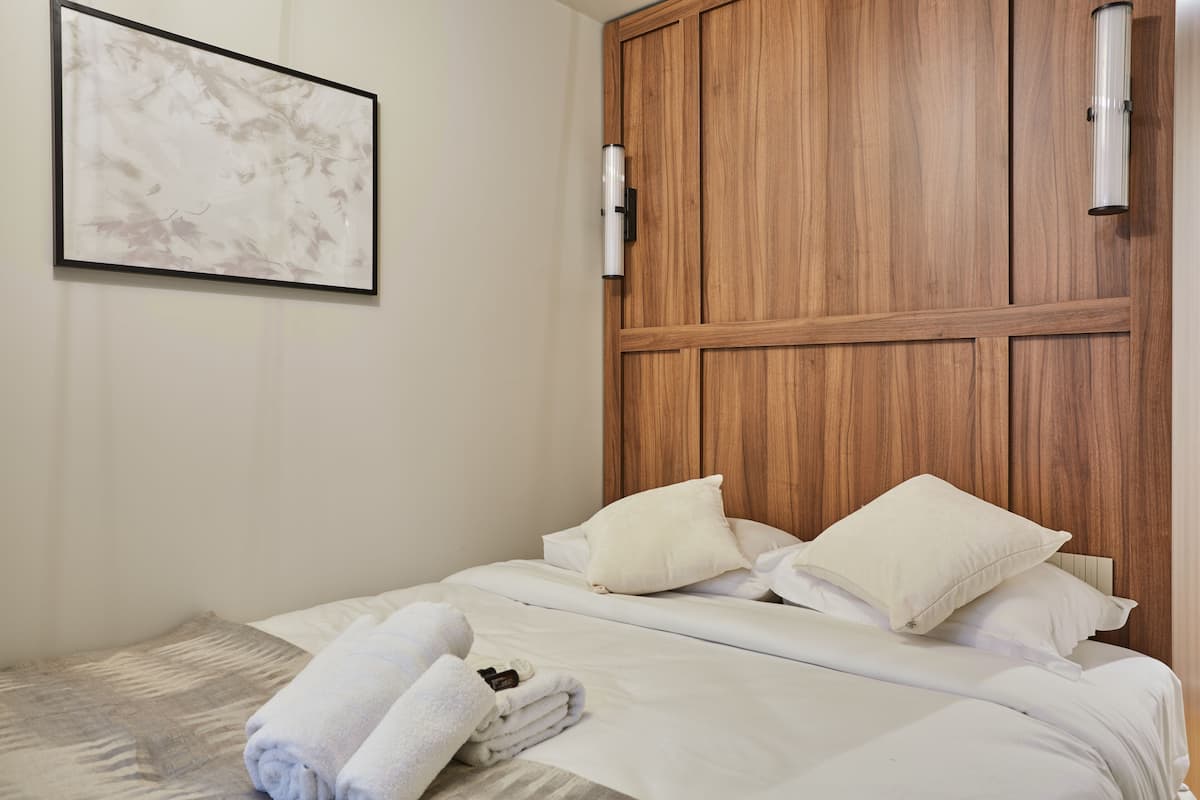 A neatly made bed with white linens and pillows, a wooden headboard, and a framed artwork on the wall.