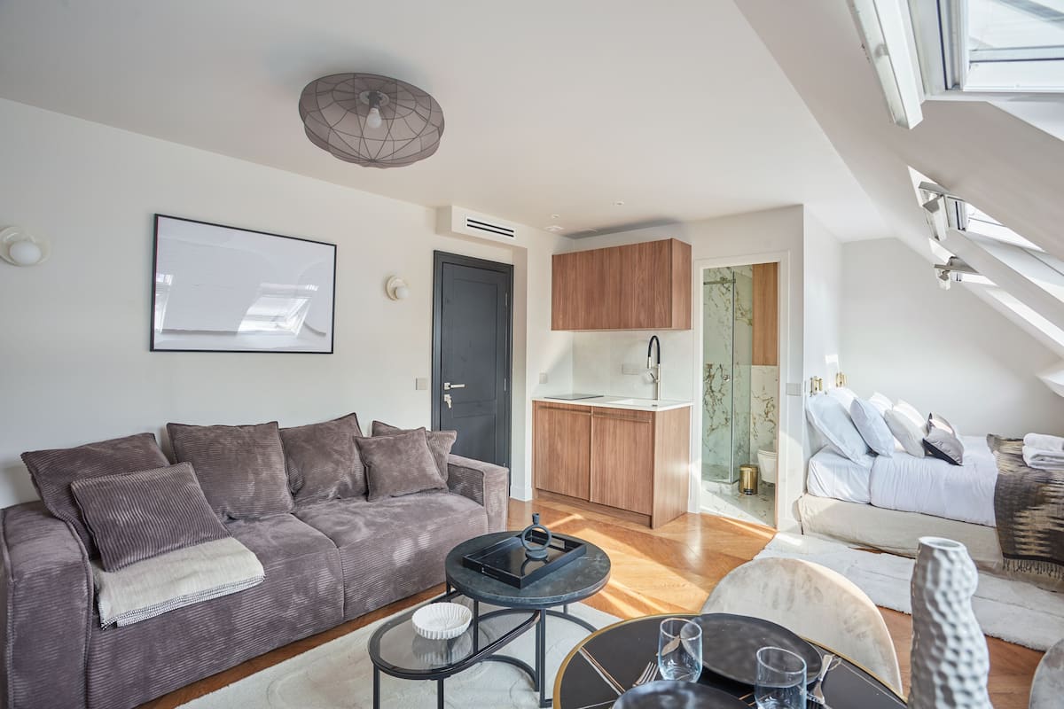 A modern living room with a grey sofa, a coffee table, and a dining area with a small table and chairs. There is a kitchen area with wooden cabinets and a window.