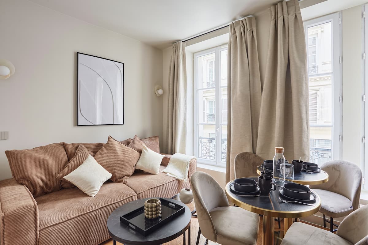 A modern living room with a brown sofa, a round dining table with chairs, and a large window with curtains.