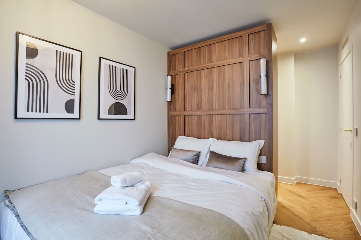 A neatly made bed with white linens and a folded towel, a wooden headboard, and two framed abstract artworks on the wall.