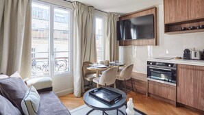 Ett kompakt vardagsrum med en soffa, ett matbord, stolar, en köksdel med en tv och ett fönster med gardiner.