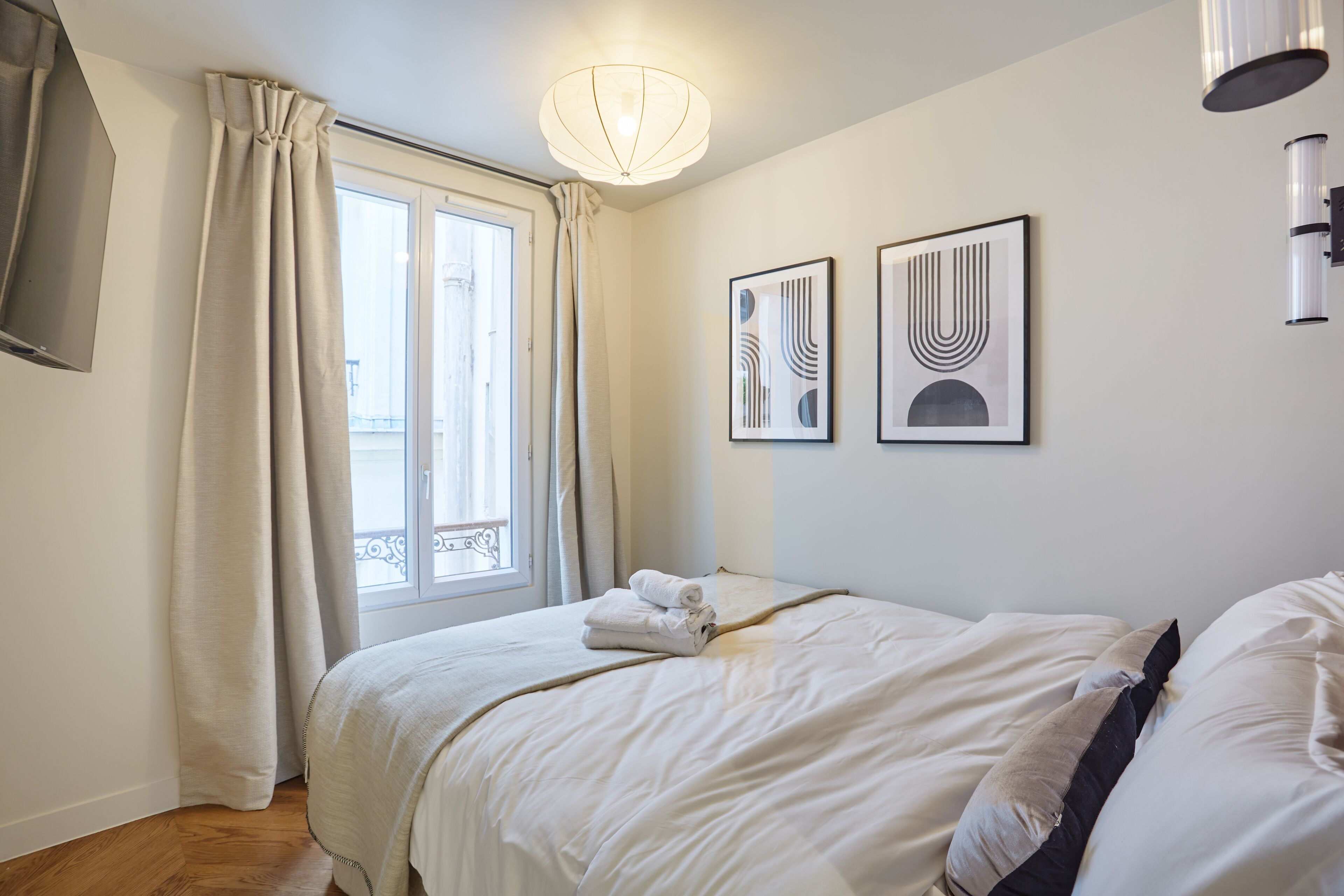 A neatly made bed with a white comforter and pillows, a window with curtains, and two framed artworks on the wall.