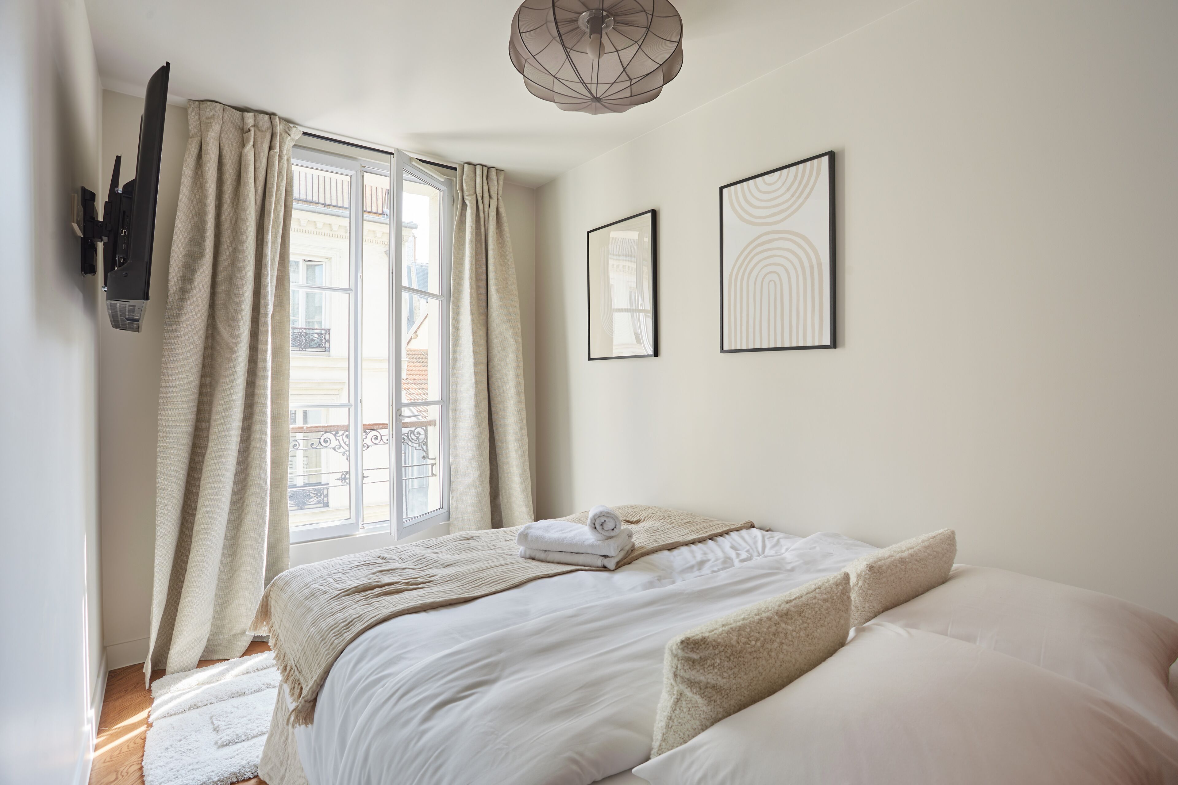 A neatly made bed with a white comforter and pillows, a window with curtains, and two framed artworks on the wall.
