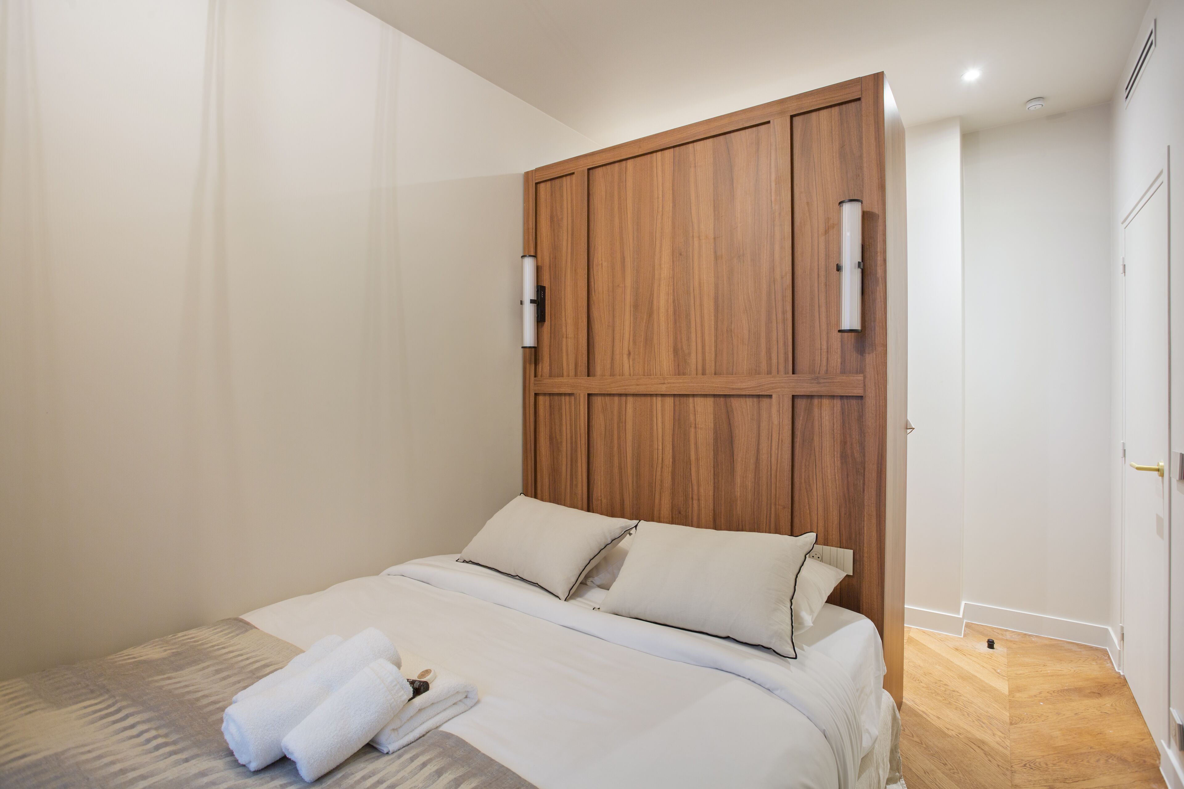 Un lit bien fait, avec du linge de lit blanc et des oreillers, une tête de lit en bois et une petite table de chevet sur laquelle est posée une serviette blanche.