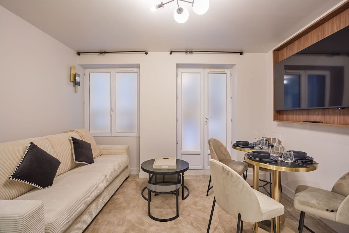 A modern living room with a beige sofa, two black cushions, a round coffee table, and a dining table with chairs.