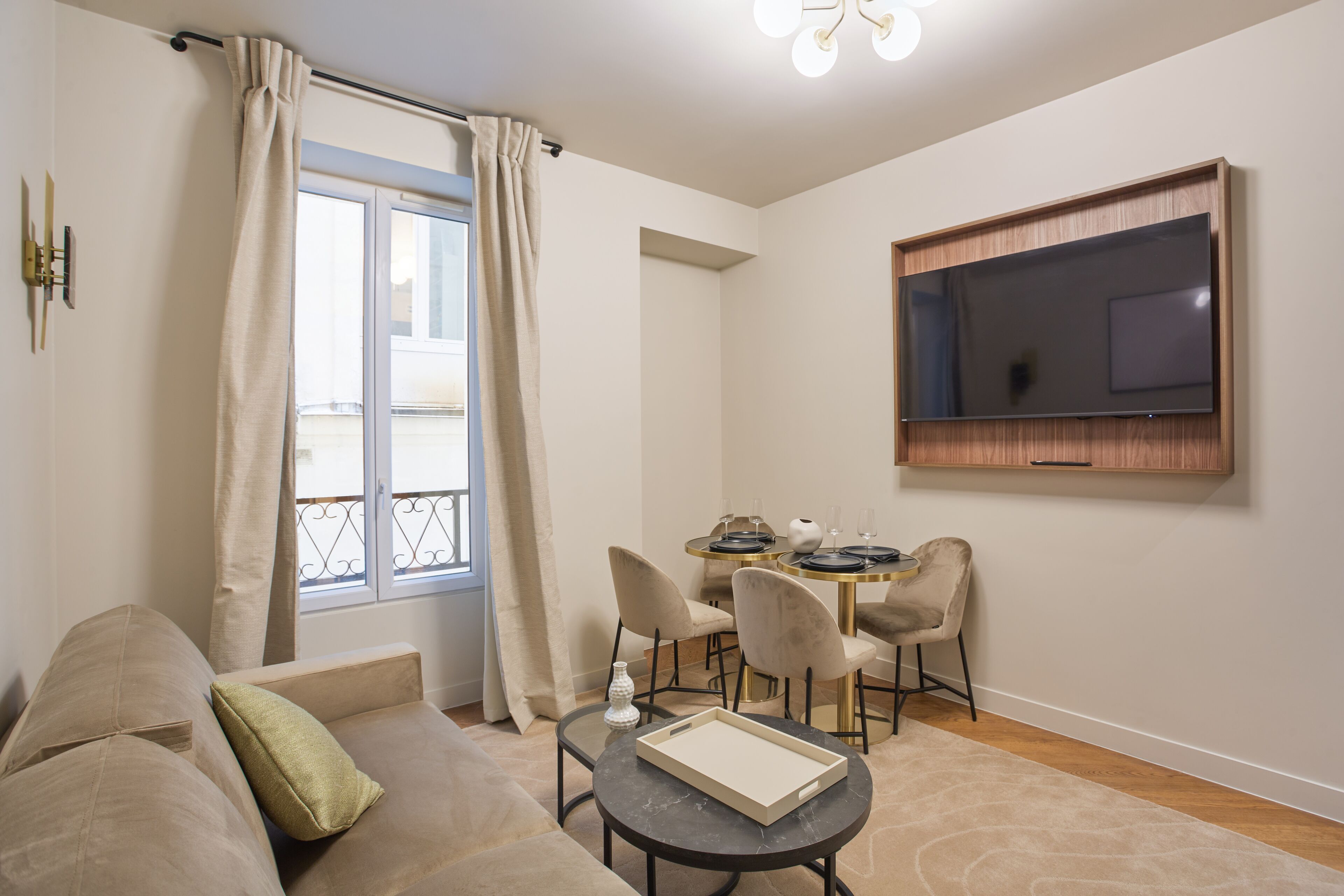 A modern living room with a sofa, a small round table with a tray, a dining area with chairs, a TV mounted on the wall, and a window with curtains.