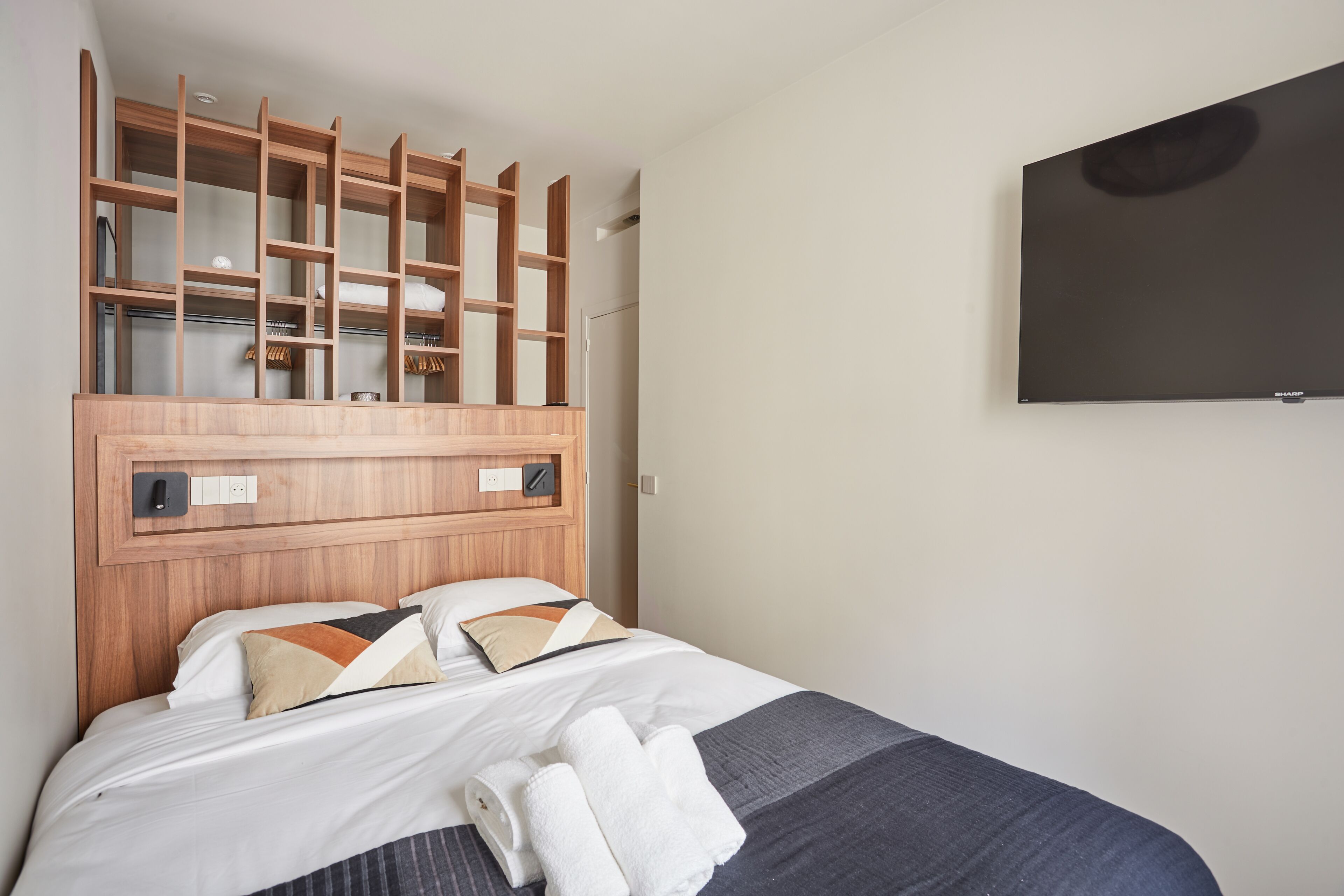 A hotel room with a wooden headboard, a bed with white linens, a flat-screen TV mounted on the wall, and a built-in closet with shelves.