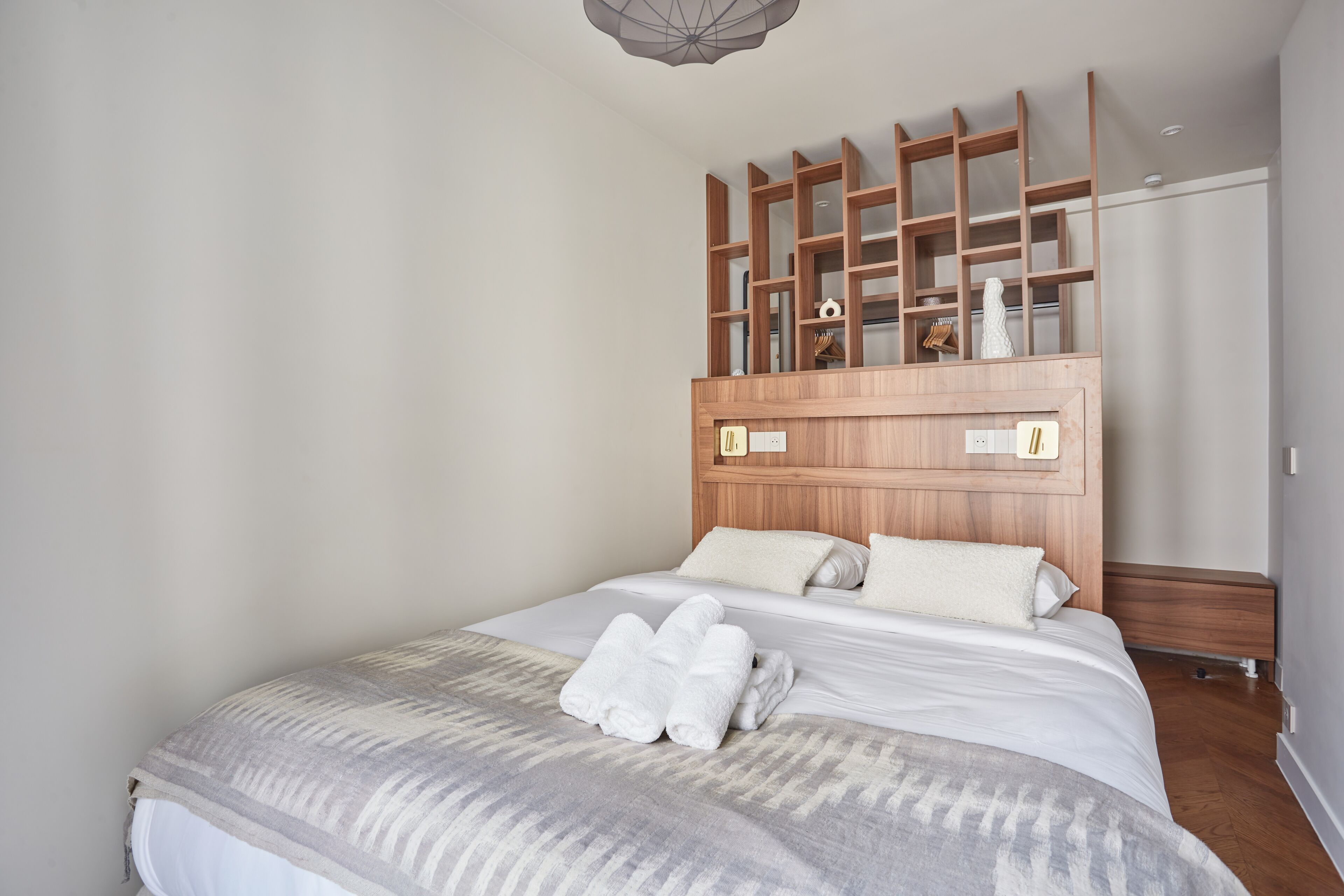 A neatly made bed with white linens and rolled towels, a wooden headboard with built-in shelves, and a simple, clean room design.