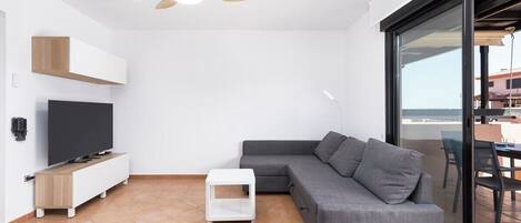 A modern living room with a grey sectional sofa, a flat-screen TV on a wooden stand, a ceiling fan, and a large sliding glass door leading to an outdoor area.