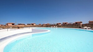 Uma grande piscina com borda curva, cercada por um muro de madeira e uma fileira de edifícios com arquitetura uniforme.
