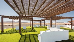 Uma área externa coberta com estrutura de pérgola, grama sintética e vasos brancos.