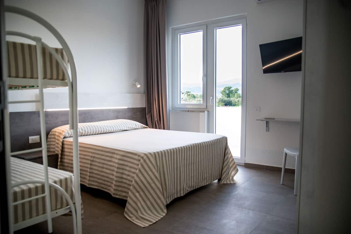 A room with a bunk bed, a single bed, a TV mounted on the wall, and a window with a view of greenery.