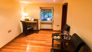 Una habitación con piso de madera, un escritorio con lámpara, una silla, una mesa de comedor con una bandeja de copas de vino y rosas, y una ventana con vista a árboles.