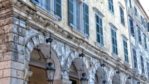Un edificio histórico con múltiples arcos y columnas, terraza con mobiliario de mimbre y una hilera de ventanas con contraventanas.