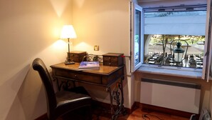 Habitación de esquina con escritorio de madera, una silla, una lámpara y una ventana que da a mesas y sillas al aire libre.