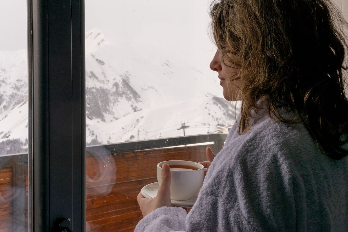 A person in a robe holding a cup, looking out of a window at a snowy mountain landscape.
