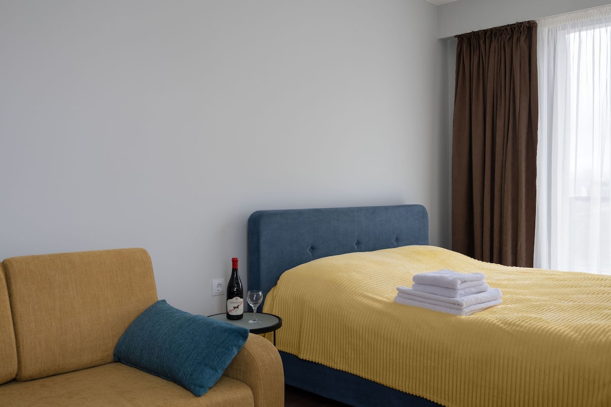A hotel room with a bed, a sofa, a small table with a bottle and glass, and a window with curtains.