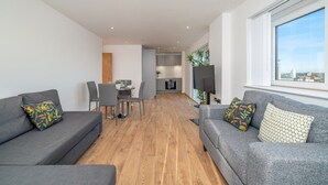A modern living room with grey sofas, a dining table, and a kitchen in the background.