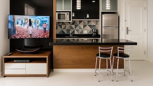 A modern kitchen with a black countertop, stainless steel appliances, and a wooden cabinet. There are two bar stools and a television mounted on a wooden cabinet. - Elegance Suites Alvorada (São Paulo)