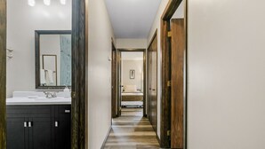 Un couloir avec une salle de bain sur la gauche, comprenant un lavabo et un miroir. Le couloir est doté de portes en bois et d’un sol carrelé.
