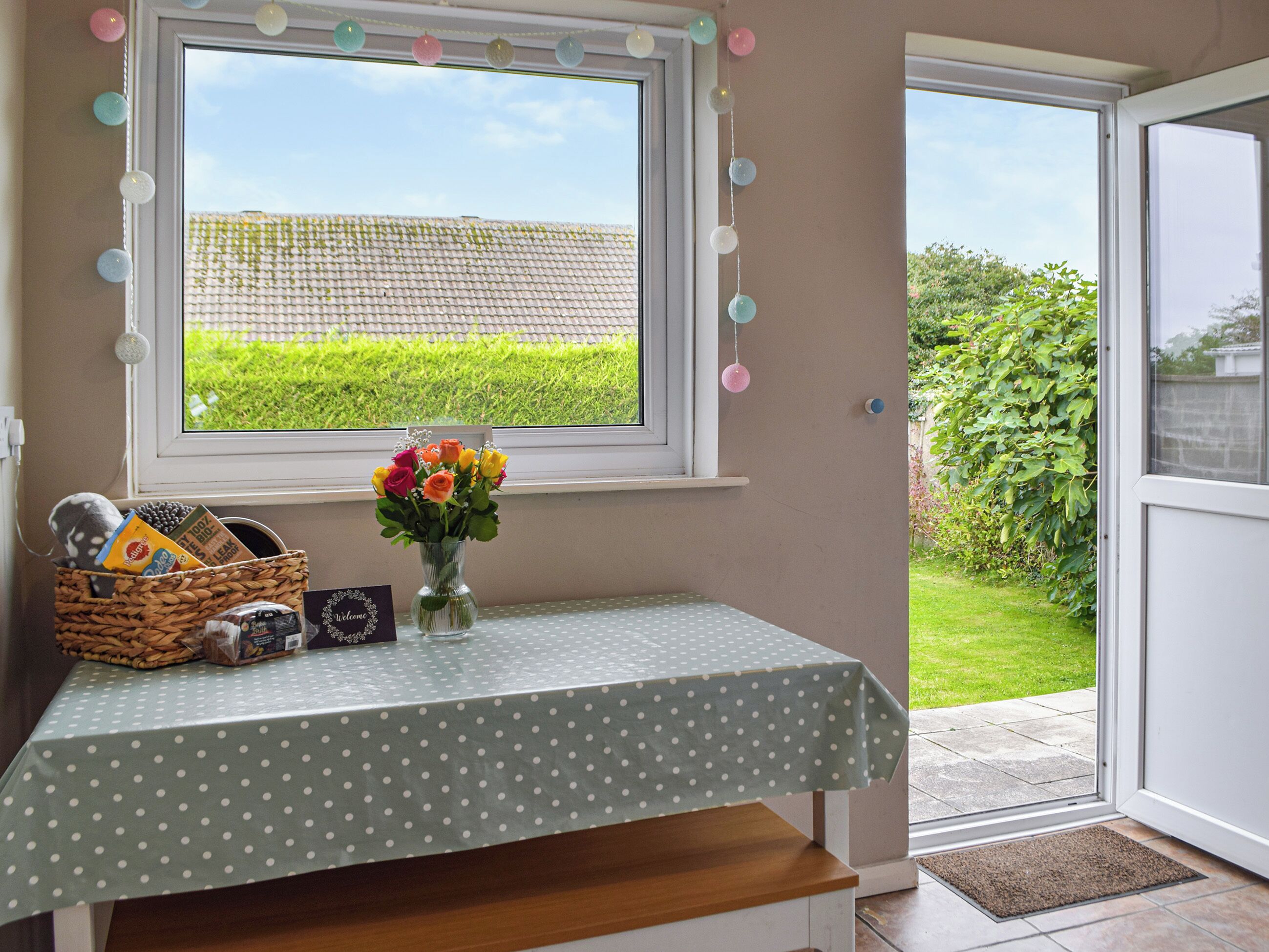 Una habitación con ventana que da a un campo verde, un mantel con lunares, un jarrón con flores y una cesta con aperitivos.