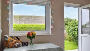 A room with a window overlooking a green field, a polka dot tablecloth, a vase with flowers, and a basket with snacks.