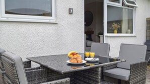 A dining area with wicker furniture, a table with croissants, fruit, and orange juice, and a view of the house.
