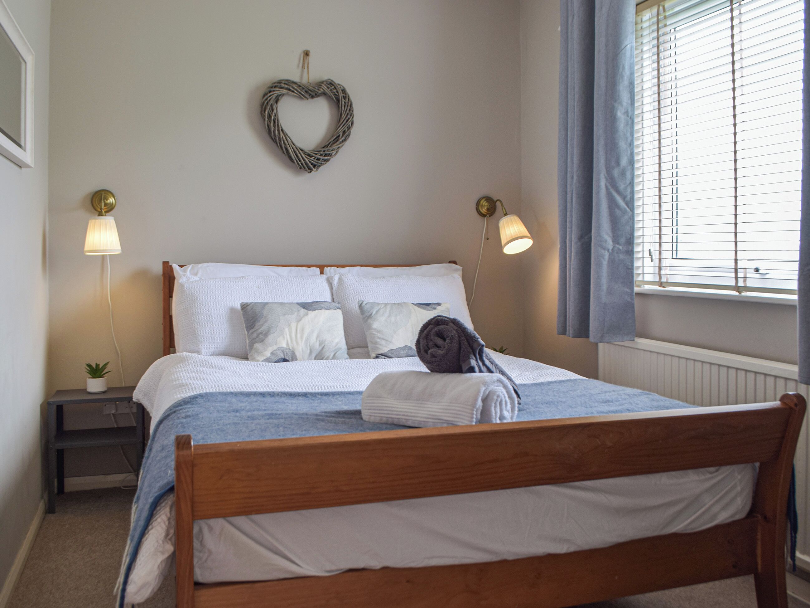 A neatly made bed with a wooden headboard, white bedding, and a rolled towel. There are two bedside lamps, a small plant, and a heart-shaped decoration on the wall.