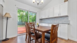 A kitchen with white cabinets, a wooden dining table with chairs, and a view through the glass door to the outdoors.