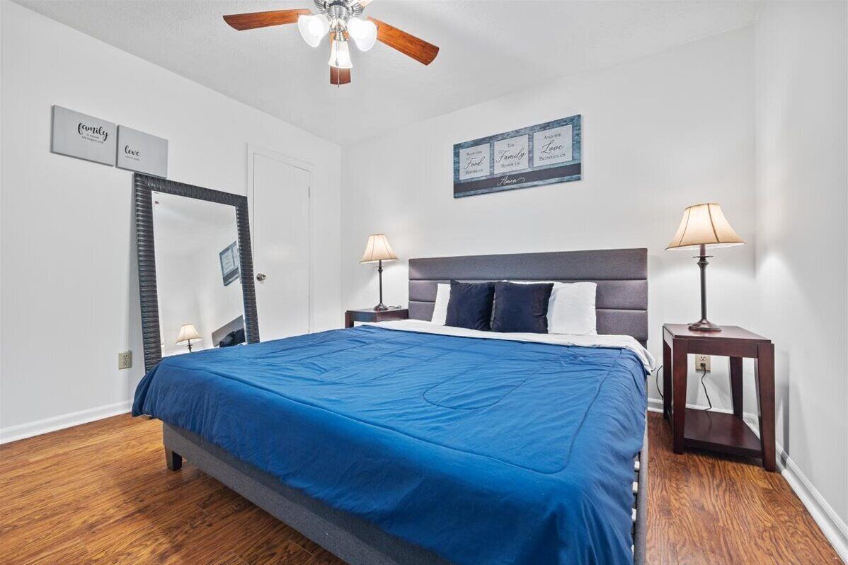 A bedroom with a bed, two bedside tables with lamps, a ceiling fan, a mirror, and wall art.