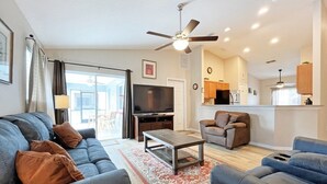 A modern living room with a sofa, armchairs, a coffee table, and a TV. There is a ceiling fan, a kitchen area, and a sliding glass door leading to an outdoor space. - Sunny pool home with private deck - great location (Davenport)