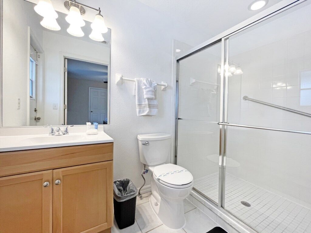 A bathroom with a white sink, mirror, and toilet. There is a glass shower enclosure and a trash can.