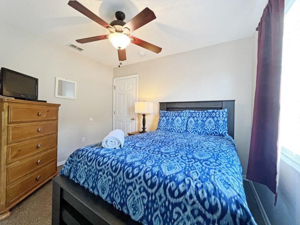 Une chambre à coucher comprenant un lit, un ventilateur de plafond, une commode, une lampe et un téléviseur.