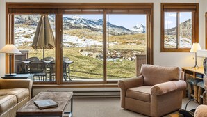 A cozy living room with a large window offering a mountain view. There is a leather sofa, a wooden coffee table, and a dining area with chairs and a table. - Condo in Snowmass Village (Snowmass Village)