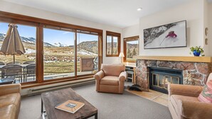 A spacious living room with a fireplace, large windows offering a mountain view, and comfortable seating. - Condo in Snowmass Village (Snowmass Village)