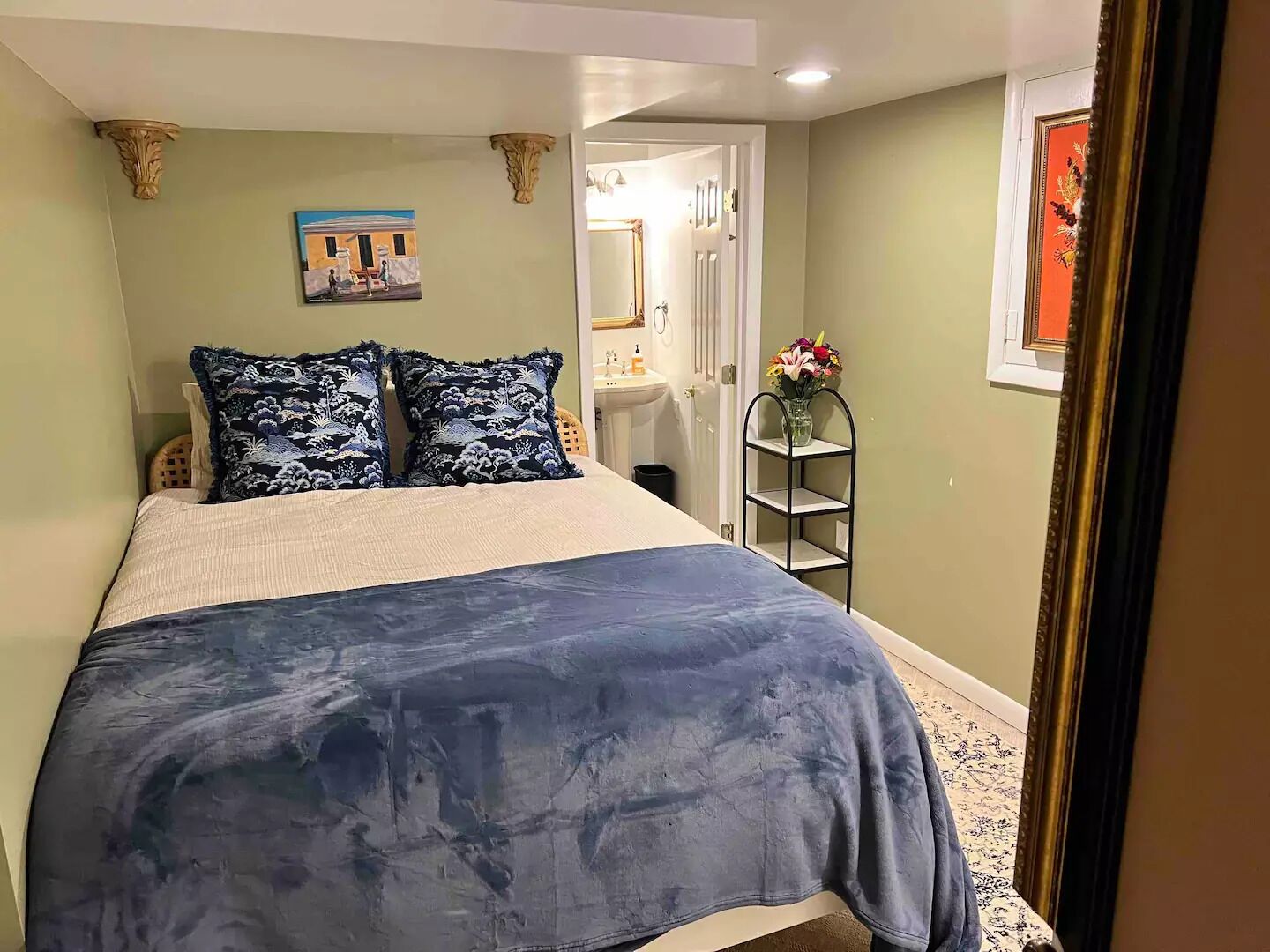 A bedroom with a bed, pillows, a painting, a mirror, a shelf with flowers, and a door.